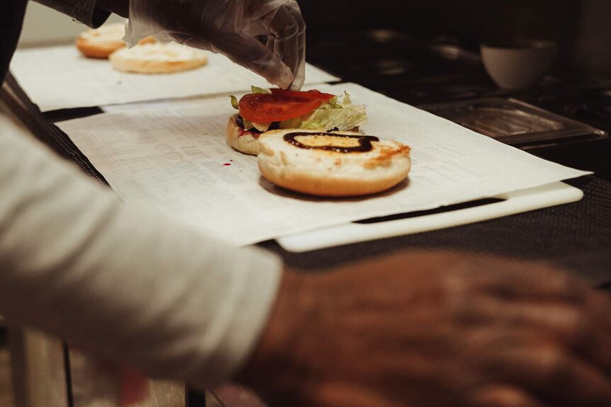 A hand in a plastic glove places a slice of tomato on an open burger bun with sauce and lettuce already on it.