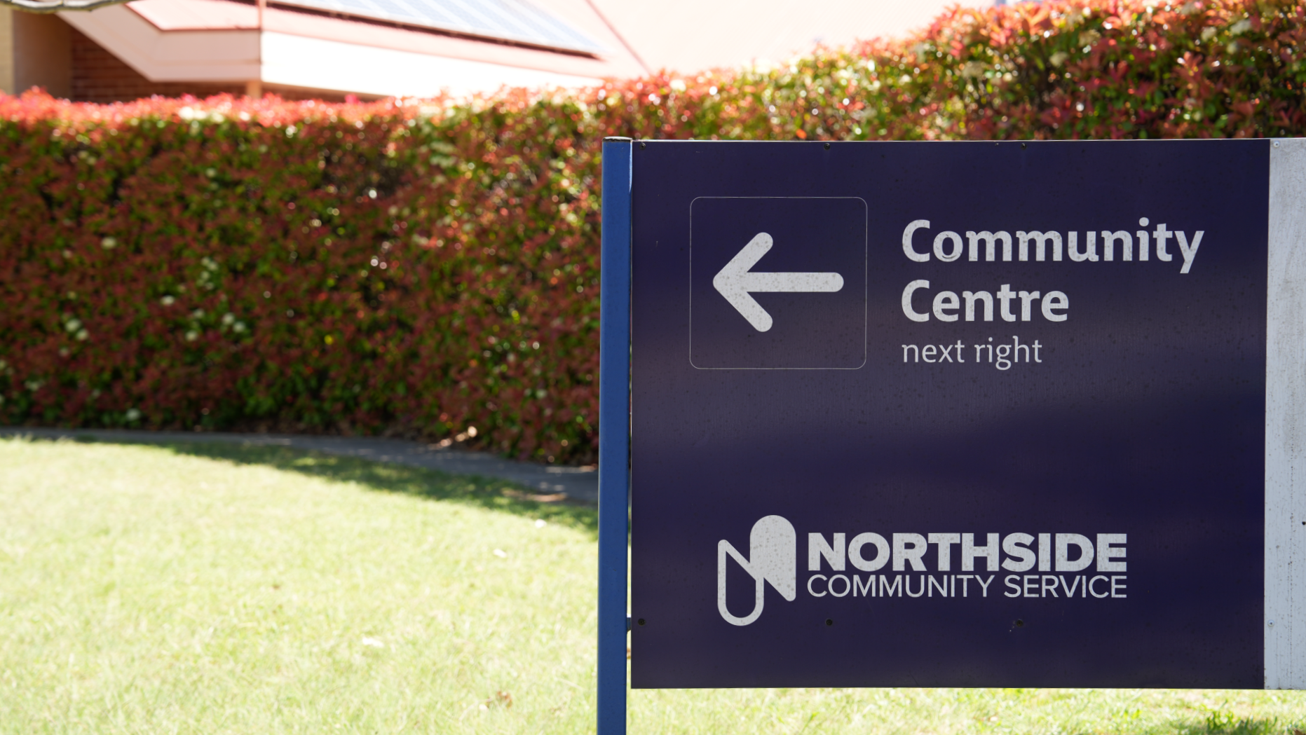 Sign saying  'Community Centre next right' and underneath says 'Northside Community Service'. Grass and hedges in background