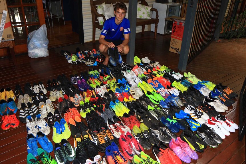 Boy kneels down beside dozens of pairs of football boots in room of home