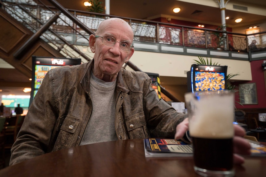 Bald man with glasses in a leather jacket sits at a high table with a pint of Guinness in front of him.
