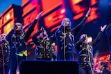 A group of young Indigenous Australian singers wear black hoodies with metallic tape on them and perform on a red stage.