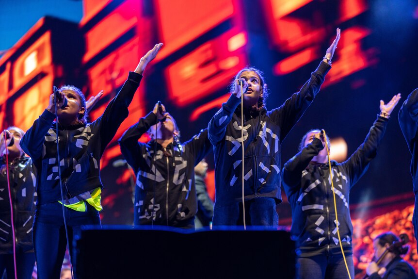A group of young Indigenous Australian singers wear black hoodies with metallic tape on them and perform on a red stage.