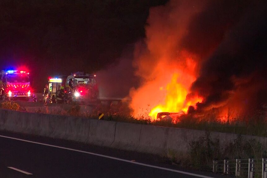 A truck on fire with emergency service vehicles closeby before dawn.