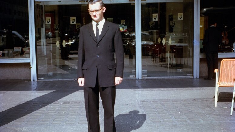 Vincent Ryan pictured in Rome in 1966 wearing a black suit and glasses, standing outside an unspecified location.