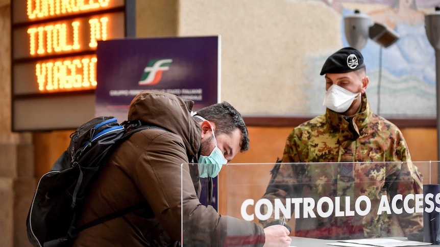 A man in a mask signs a document at a railway station watched by a solider in mask.