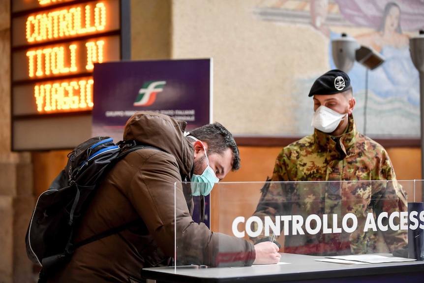 A man in a mask signs a document at a railway station watched by a solider in mask.