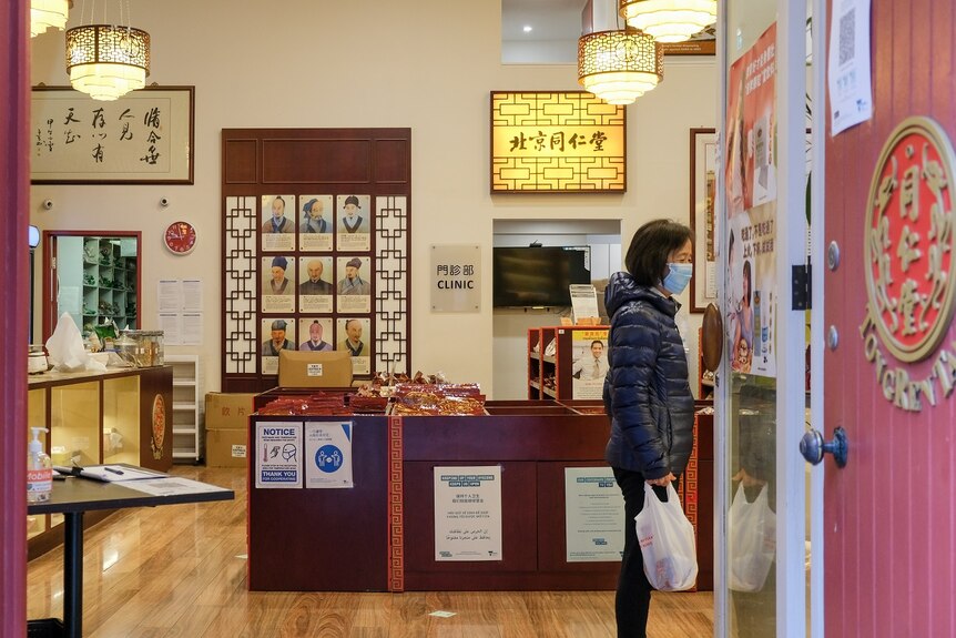 A woman wearing a mask stands inside a clinic filled with Chinese language posters.