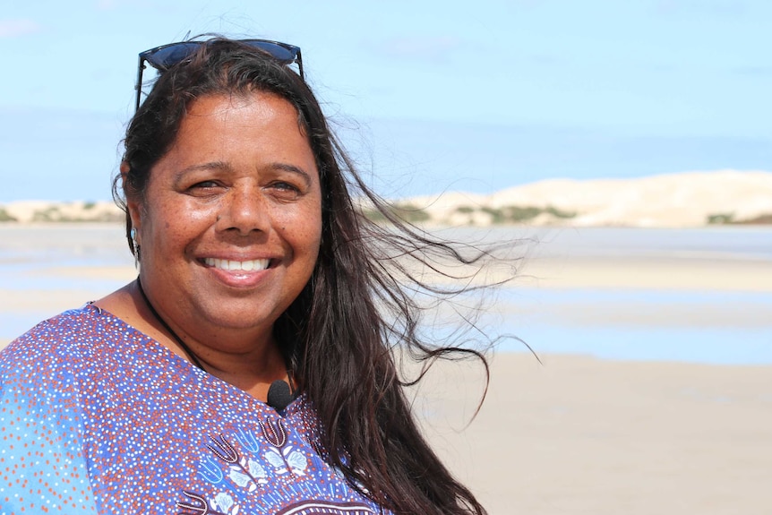 On left Woman smiling looking at camera, sunny sandy beach in background with sandhills