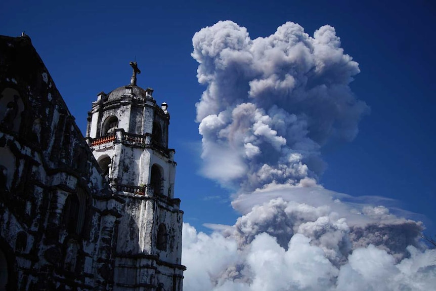 Wide shot of an old building in the foreground with a plume of volcano smoke in the background.