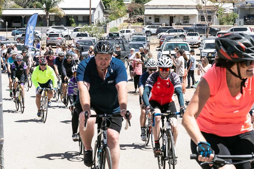 A throng of cyclists pedal down the street of a country town.