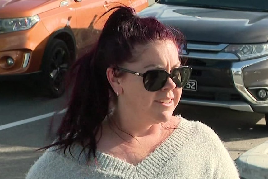 A lady with her long dark hair tied in a pony tail wears a beige jumper as she is interviewed in a parking lot