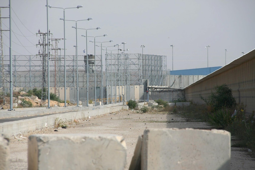 A wide photo of large concrete blocks and street lights lining a large outdoor concrete floor in front of a tall border wall.