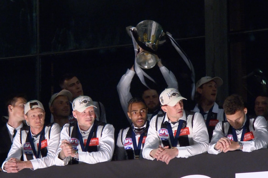 Collingwood players wearing their black and white guernseys and premiership medals stand on a balcony holding up the cup.