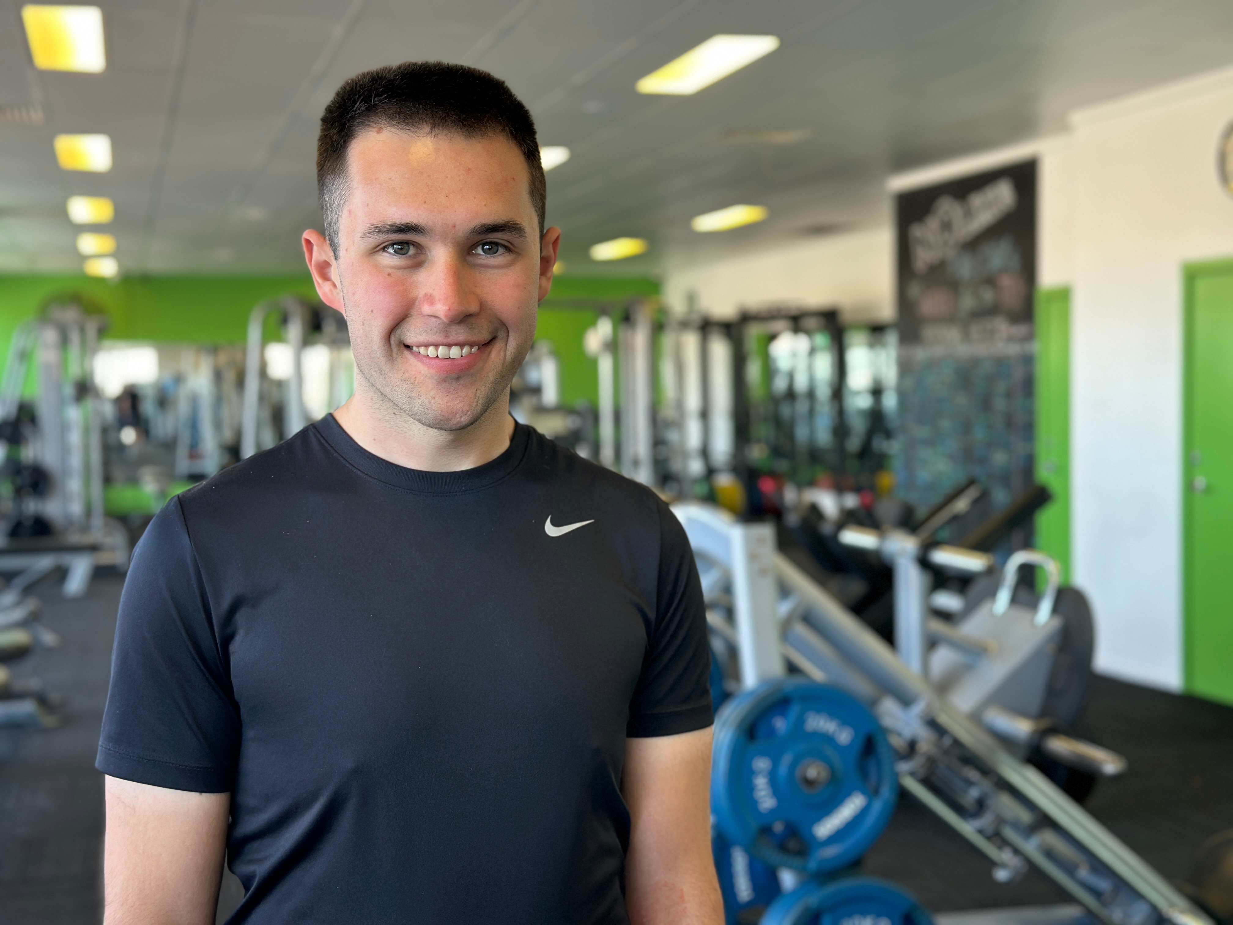 A man in a black t-shirt and black hair smiles at the camera while standing in front of a camera