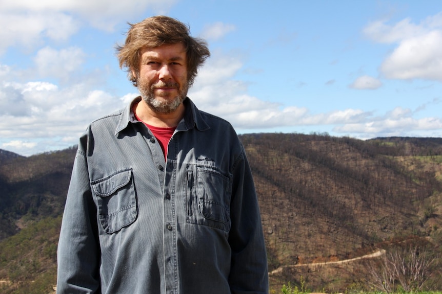 A man stands with burnt out hill and Wombeyan Caves Road in the background.