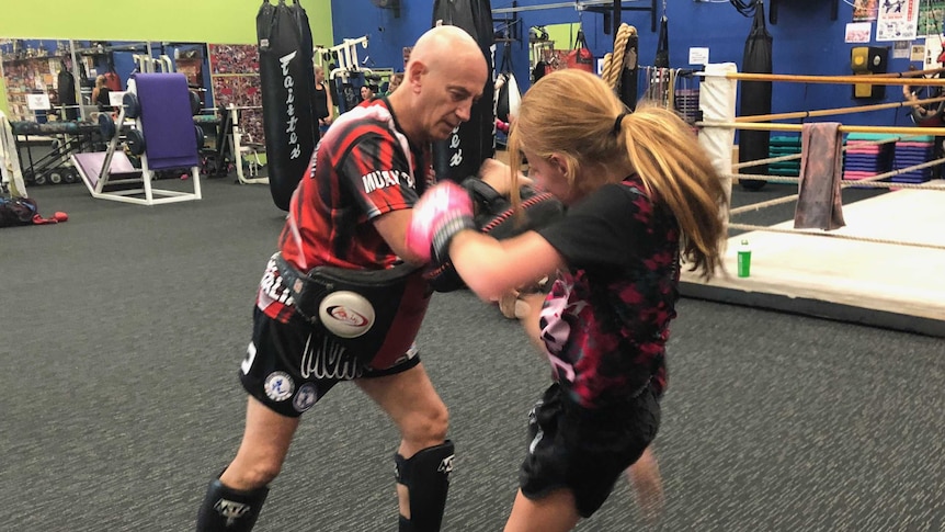 Coach Joe Hilton holds pads for young girl to use her knees in thai boxing gym