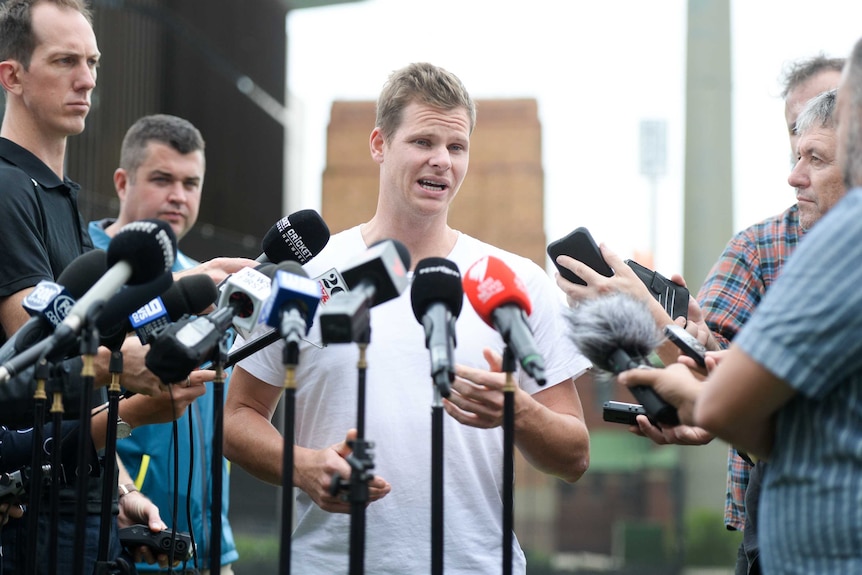 Man speaks to the media, surrounded by microphones.