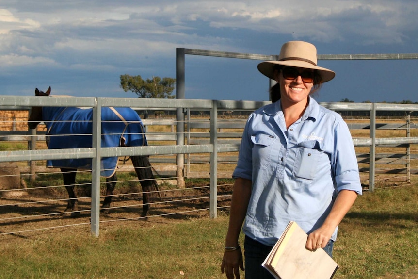 Wearing a wide-brimmed hat and sunglasses, Kelly Ostwald stands near a horse enclosure.