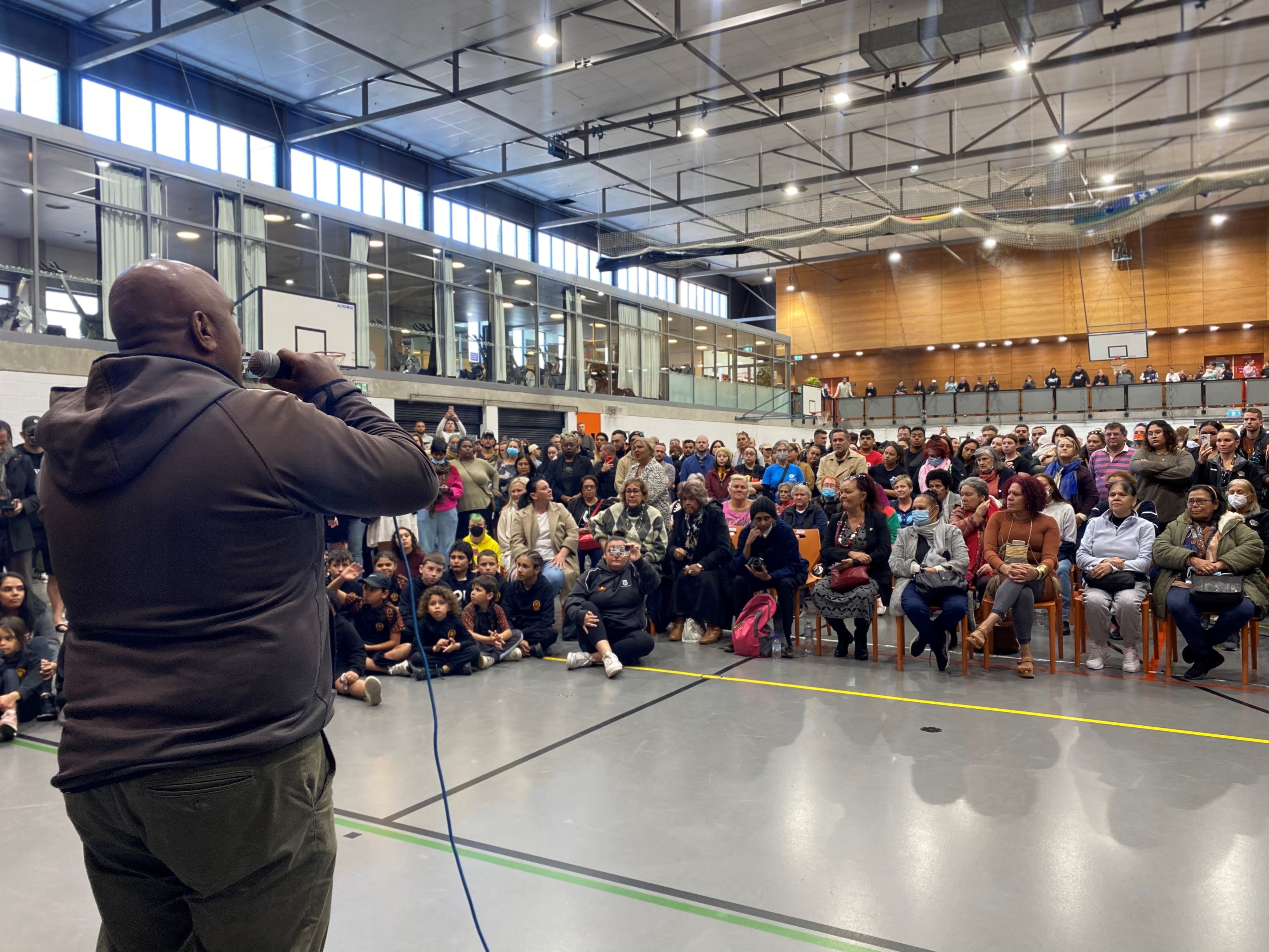 A man with his back to camera holds a microphone as he speaks to a crowd of people