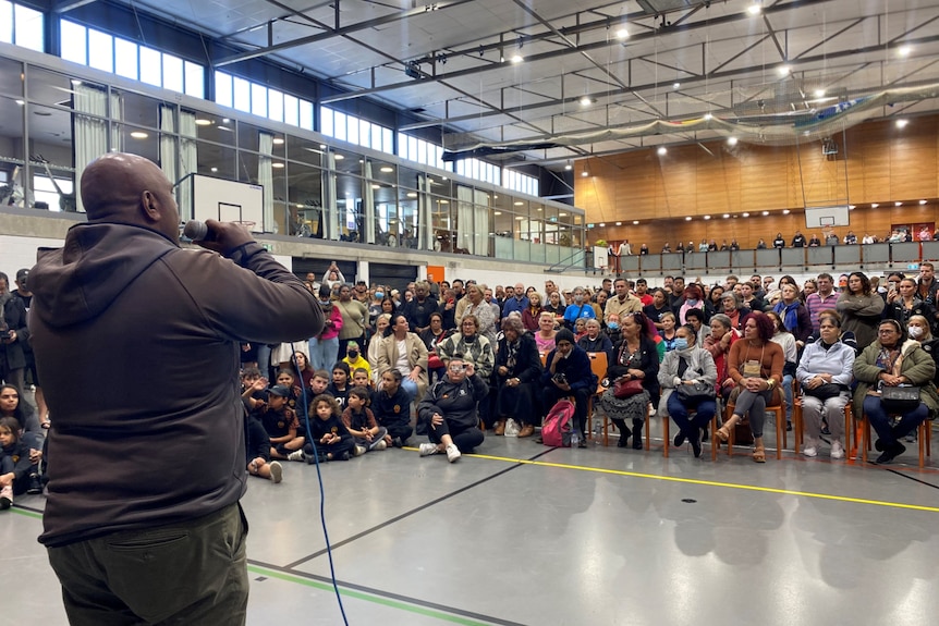 A man with his back to camera holds a microphone as he speaks to a crowd of people