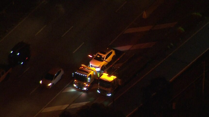 A white car on the back of a tow truck. The scene is bathed in yellow light from the tow truck light. Photo taken from above.