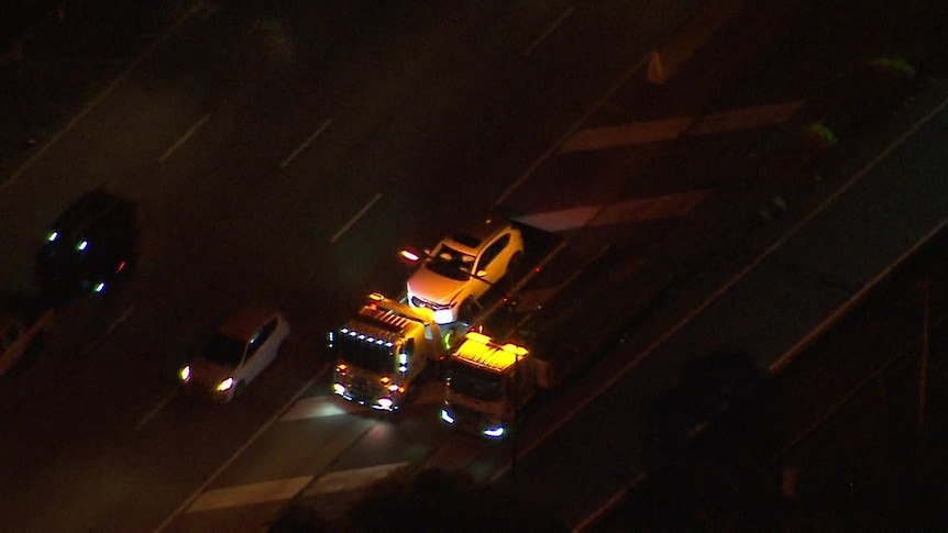 A white car on the back of a tow truck. The scene is bathed in yellow light from the tow truck light. Photo taken from above.