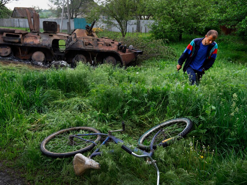 A man is a blue t-shirt walks through a field in front of a burnt-out tank