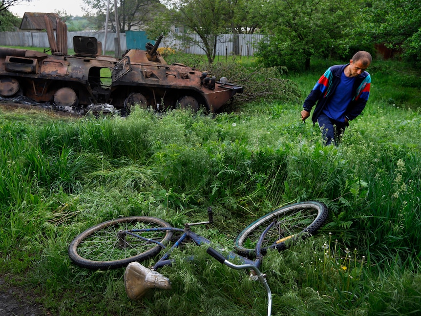 A man is a blue t-shirt walks through a field in front of a burnt-out tank