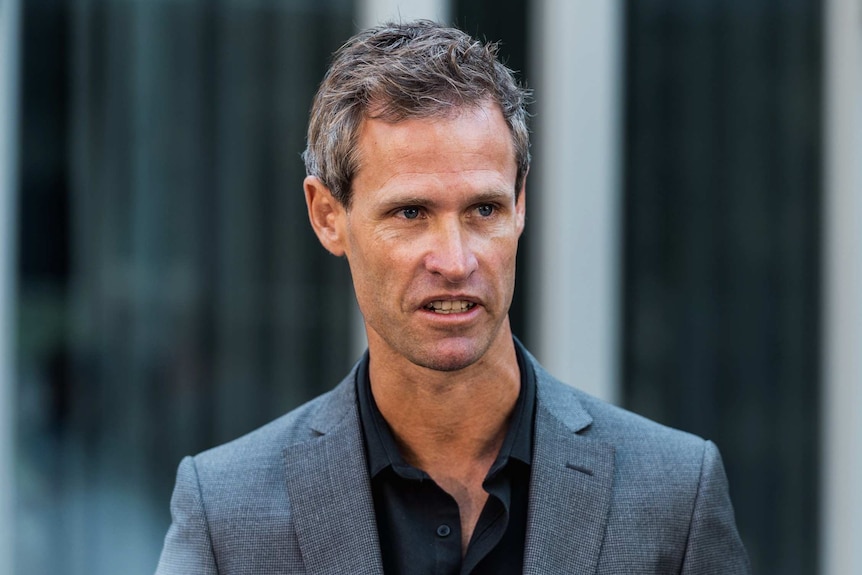 A man wearing a black shirt with open neck and a grey suit stands outside a WA court.