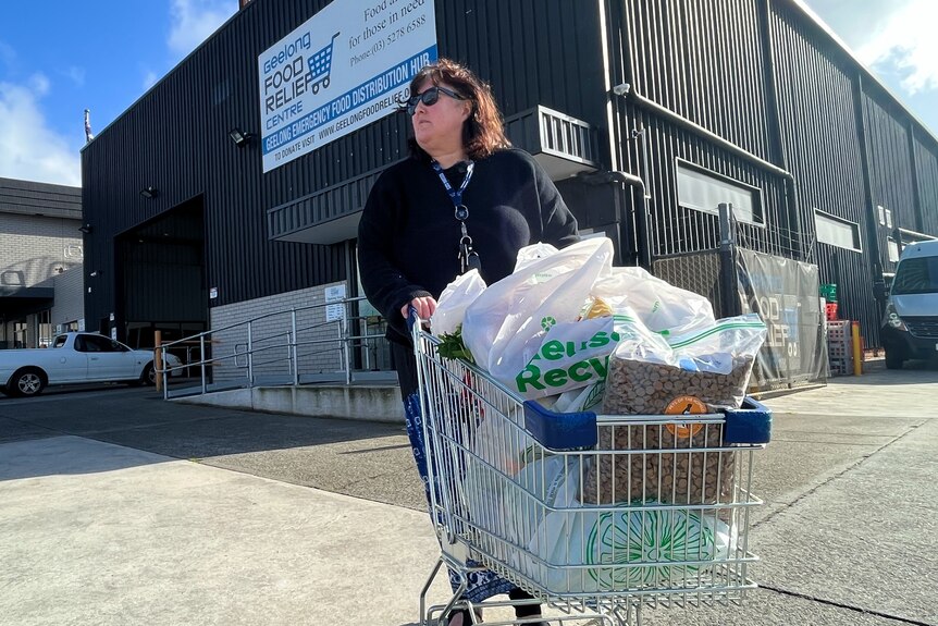 Mandy is in a car park with a shopping trolley full of plastic bags. She stands in front of Geelong Food Relief's large building