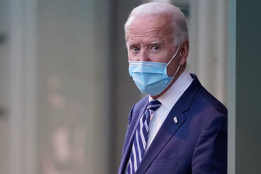 Joe Biden in a blue suit with a blue face mask walks outside