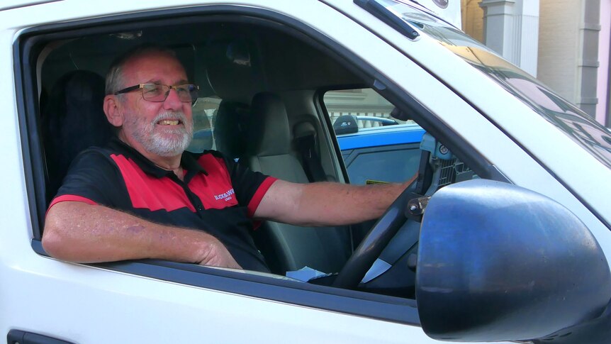 A man in a red and black shirt sitting in a taxi van with his hand on the steering wheel and looking out.