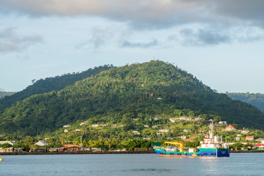 Apia Harbor in Samoa, with a large boat in the foreground and a large hill covered in trees in the background.