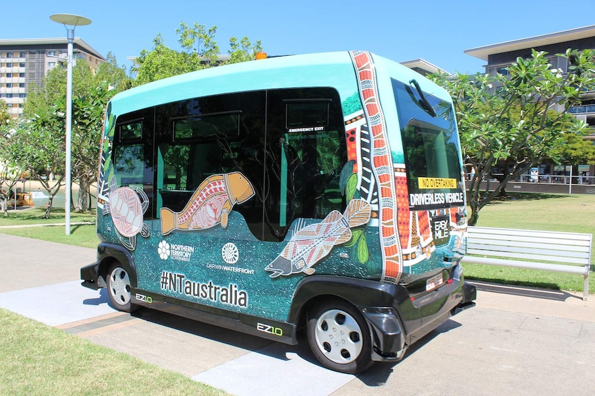 A small passenger bus decorated with Aboriginal artwork drives along a footpath in an open parkland.