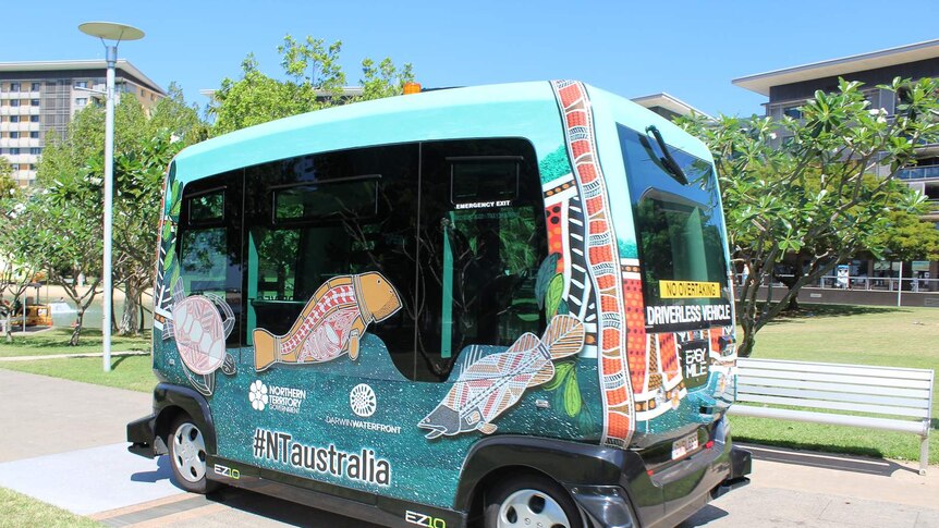 A small passenger bus decorated with Aboriginal artwork drives along a footpath in an open parkland.