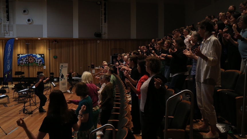 Singers stand in raked auditorium seating with their arms outstretched. Katherine Feeney demonstrates out the front.