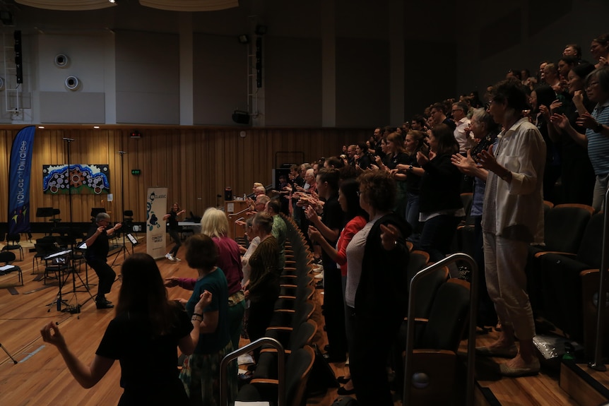 Singers stand in raked auditorium seating with their arms outstretched. Katherine Feeney demonstrates out the front.