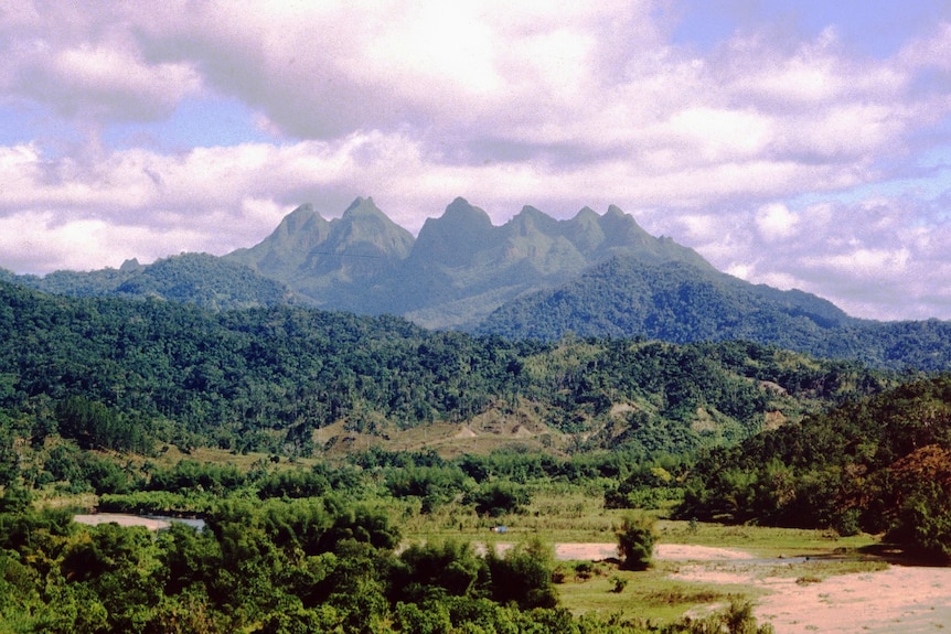A landscape showing mountains in the background, trees, shrubs, land in the foreground, clouds touching the peaks.