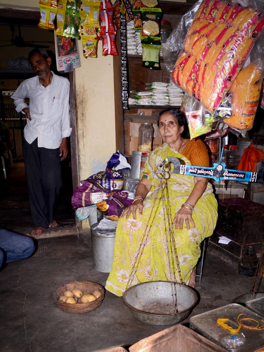 A woman wearing a yellow sari decoratid with flowers sits on a bench in a small room filled with items.