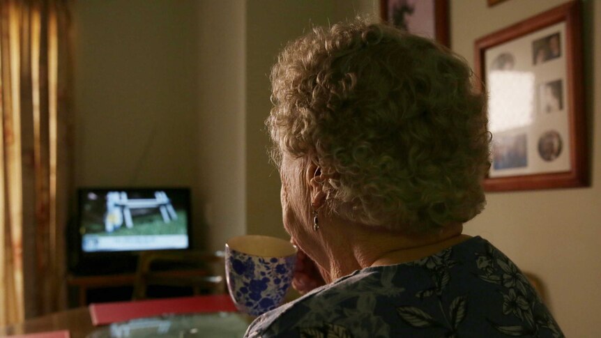 An elderly woman is pictured from behind, sipping a cup of tea while watching television. The room is dark.