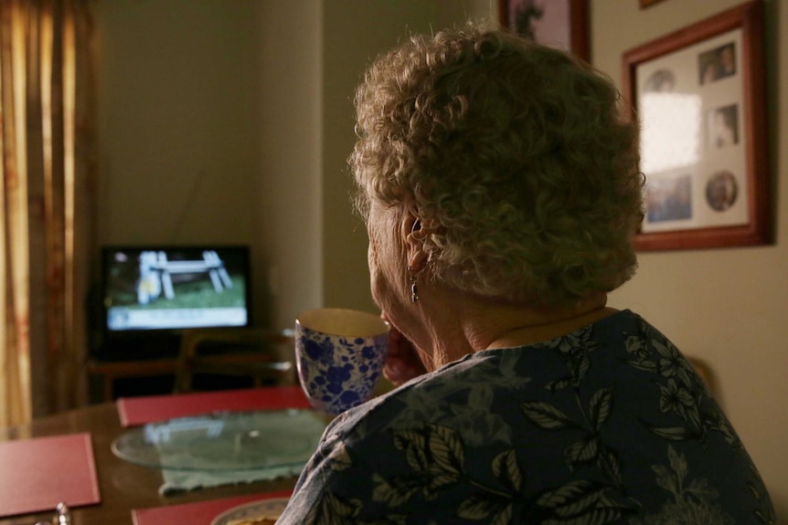 An elderly woman is pictured from behind, sipping a cup of tea while watching television. The room is dark.