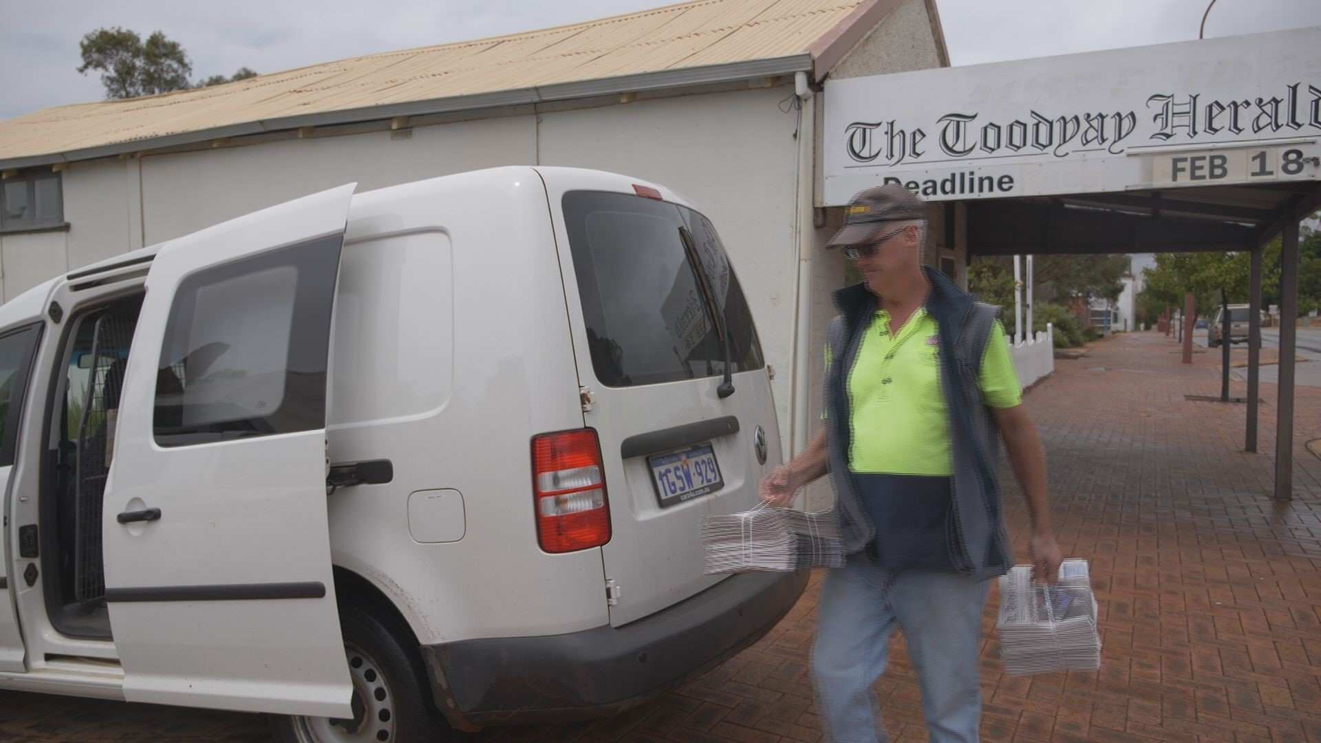 A man in a fluorescent top loads newspapers into a van outside a newspaper office.
