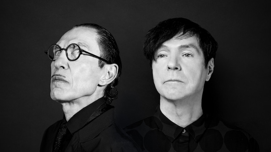 Black and white image of Ron Mael with head to the side and severe expression and a nonplussed Russell Mael