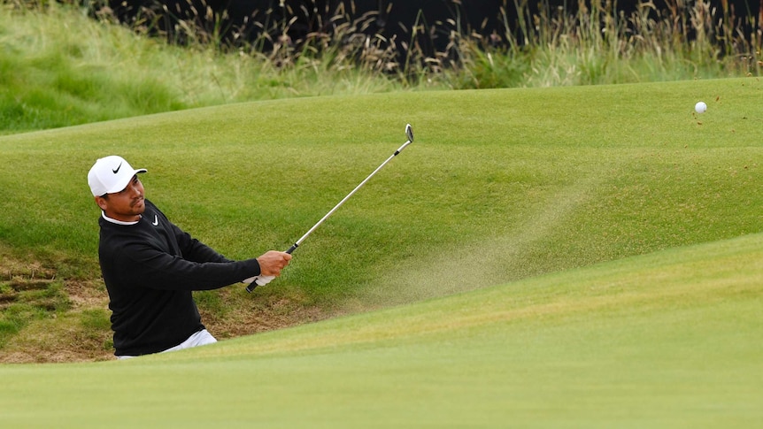 Sand flies behind the ball as Jason Day - partially obscured while standing in a bunker - completes a casual shot.