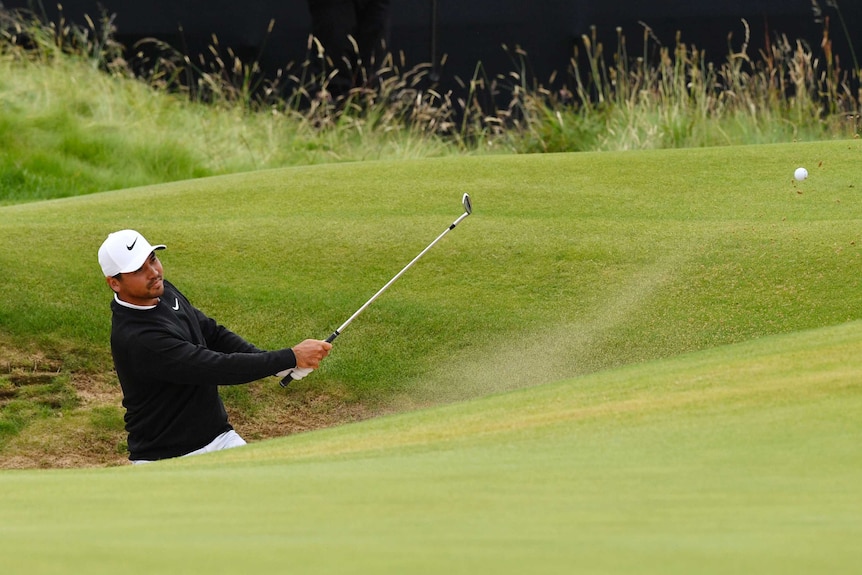 Sand flies behind the ball as Jason Day - partially obscured while standing in a bunker - completes a casual shot.