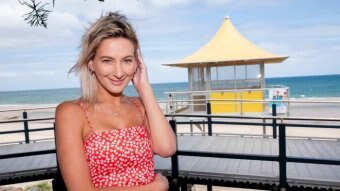 Emmah Money smiles at the camera standing at the beach, a lifeguard hut behind her.