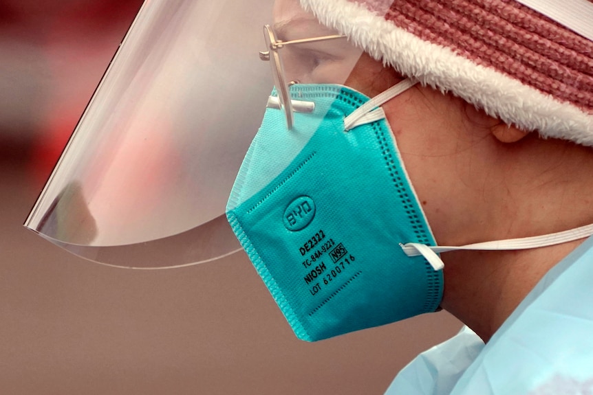 A woman shot in profile wears a clear face shield and a bright blue N95 mask