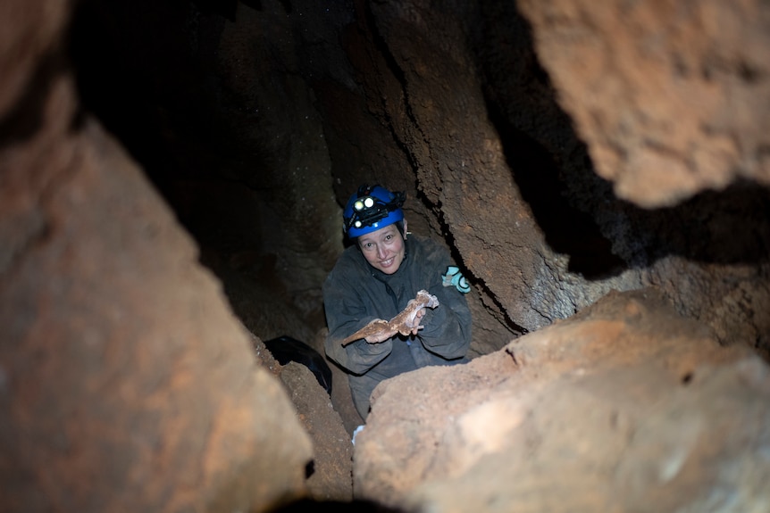 A woman in caving gear holds up a fossil and smiles