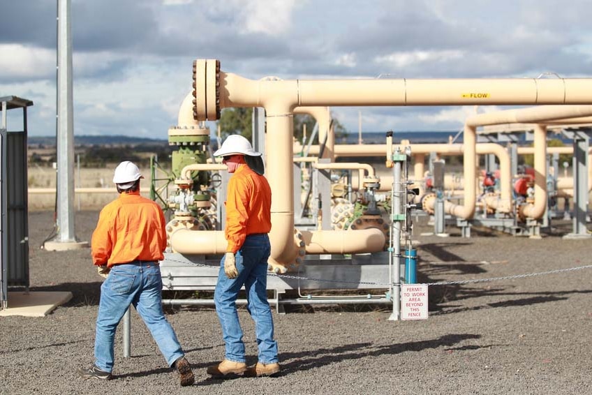 Two workers in hard hats, bright orange shirts and blue jeans walk past a gas well.