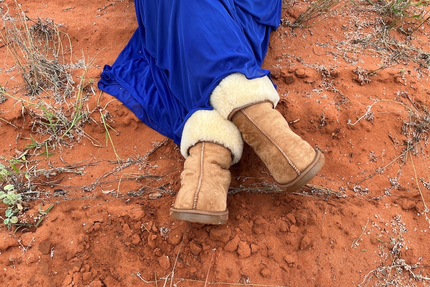 Legs crossed sitting in red sand wearing ugg boots and a blue dress. 